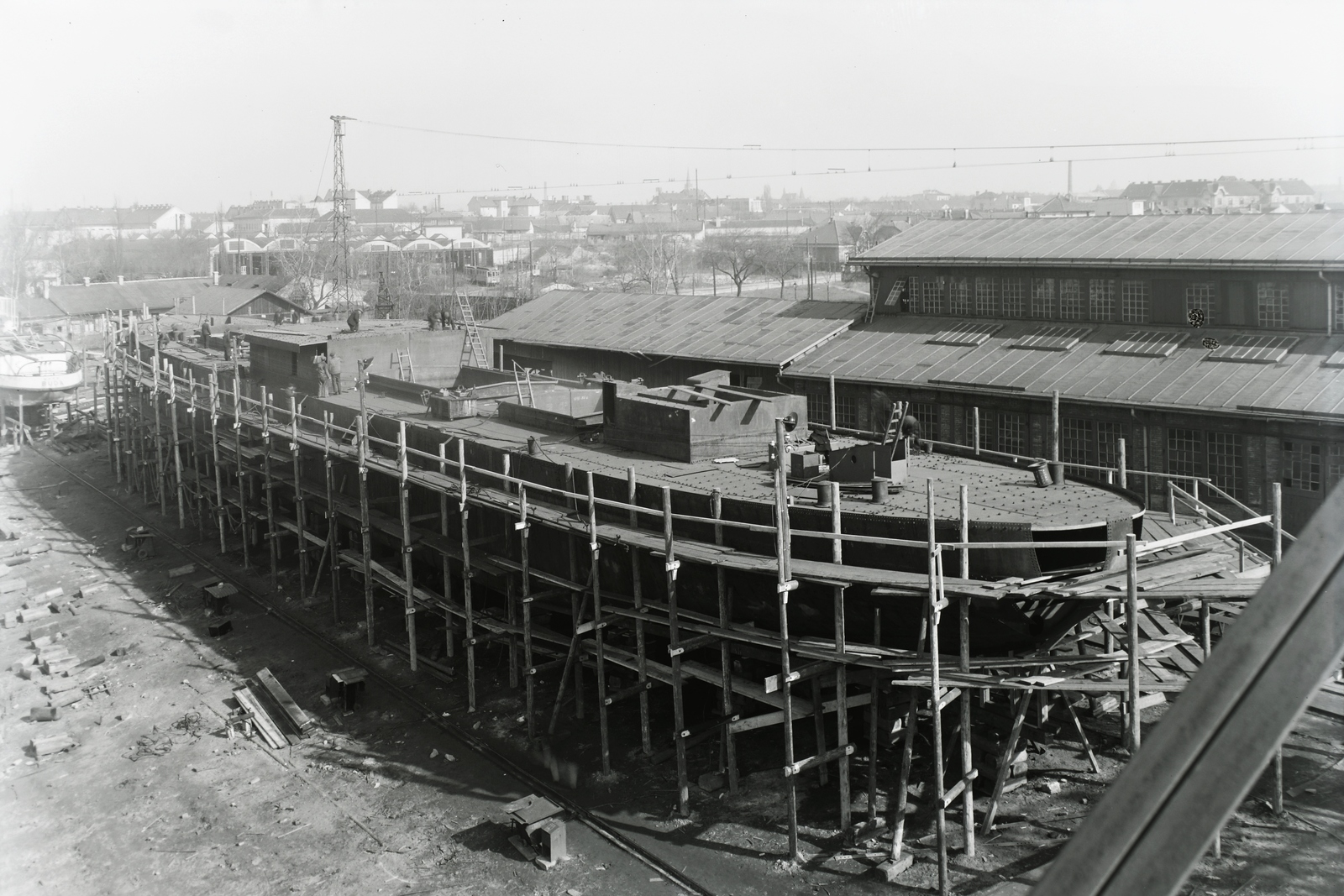 Magyarország, Budapest XIII., Újpesti-öböl, Ganz és Tsa. Villamossági-, Gép-, Waggon- és Hajógyár Rt. A sólyán az épülő DTRT Kassa (1939) Duna-tengerjáró áruszállító hajó. Háttérben a Váci úton az Újpest kocsiszín látszik. Leltári jelzet: MMKM TFGY 2017.1.389., 1939, Magyar Műszaki és Közlekedési Múzeum / Történeti Fényképek Gyűjteménye / Ganz gyűjtemény, Budapest, Fortepan #122705