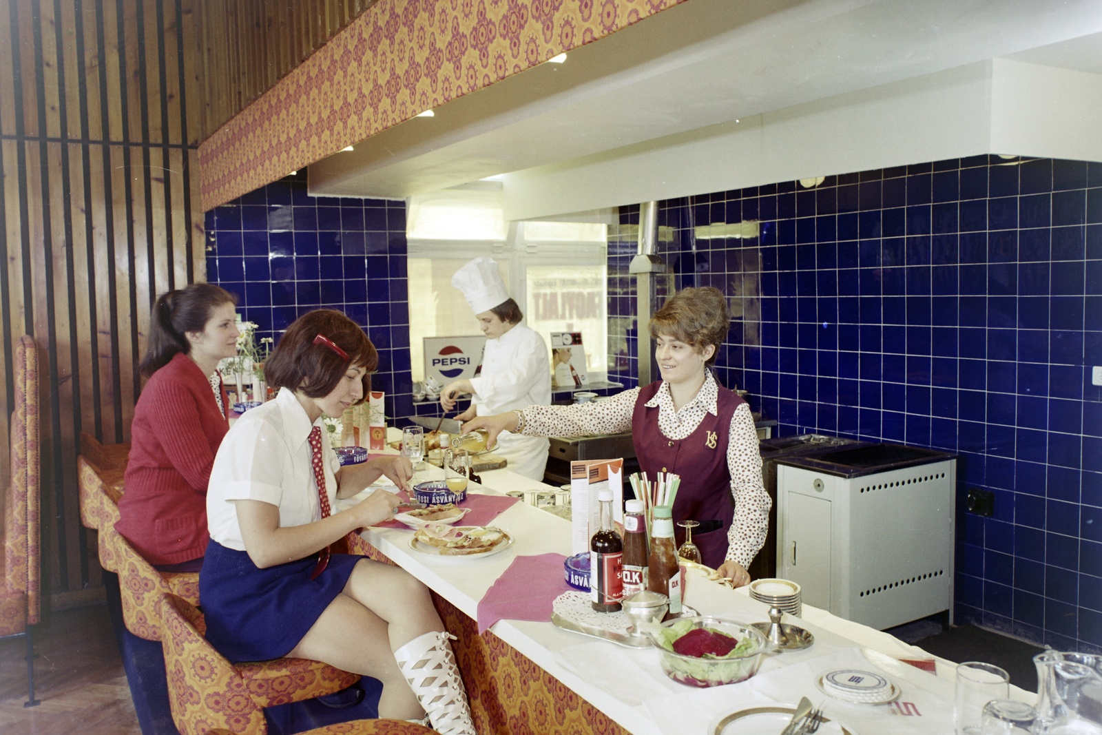 Magyarország, Budapest XI., Tas vezér utca, a Sport (később Flamenco) szálló eszpresszója., 1973, Bauer Sándor, nyakkendő, színes, egyenruha, étkezés, mosoly, csésze, pohár, jelvény, főzés, asztalterítő, hamutartó, kötény, hajviselet, étlap, szoknya, csempe, pult, szendvics, gáztűzhely, lambéria, tányér, evőeszköz, konnektor, bárszék, tükröződés, szagelszívó, bárpult, kukta, Budapest, evőeszközkészlet, uzsonna, blúz, kardigán, magasszárú cipő, lófarok, söralátét, Fésű, itallap, szívószál, Pepsi-márka, ketchup, saláta, tál, Fortepan #126590