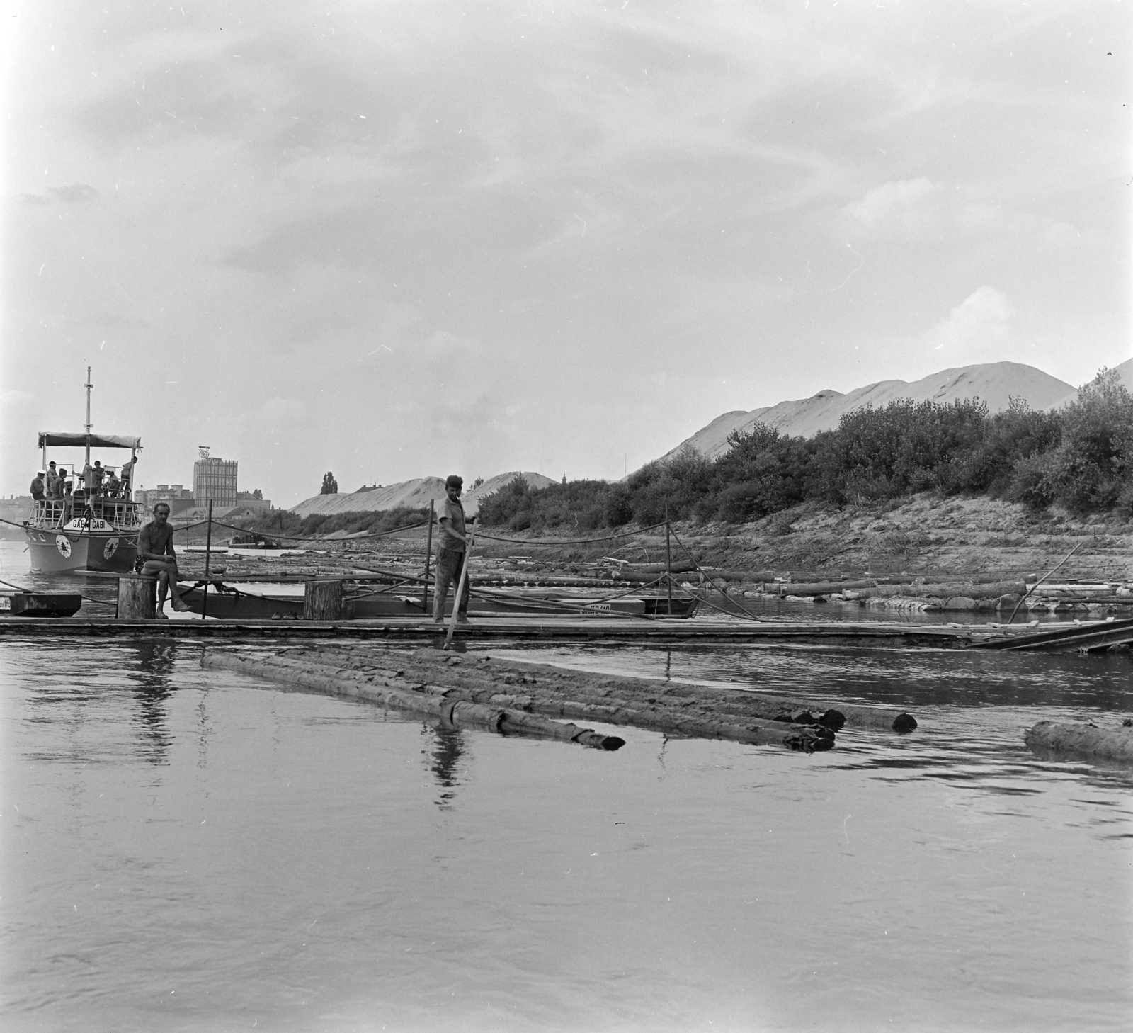 Hungary, Szeged, Tisza-part, távolban a Pick szalámigyár. A Tiszán Tuzsérról Szegedre, az ERDÉRT vállalat telepétől a fafeldolgozó üzemekhez leúsztatott farönkök. Balra az ERDÉRT vállalat Gabi nevű rönkvontató hajója., 1967, Szalay Zoltán, timber rafting, Fortepan #138017