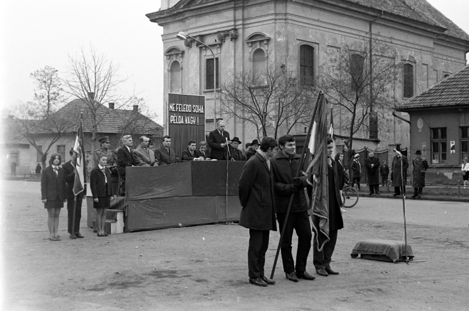 Hungary, Monor, Kossuth Lajos utca, május 1-i ünnepség a megyei pártbizottság (később könyvtár) előtt, a szovjet hősi emlékműnél. Háttérben a katolikus templom, 1968, Péterffy István, church, flag, microphone, stand, slogan, winners' podium, Fortepan #141685