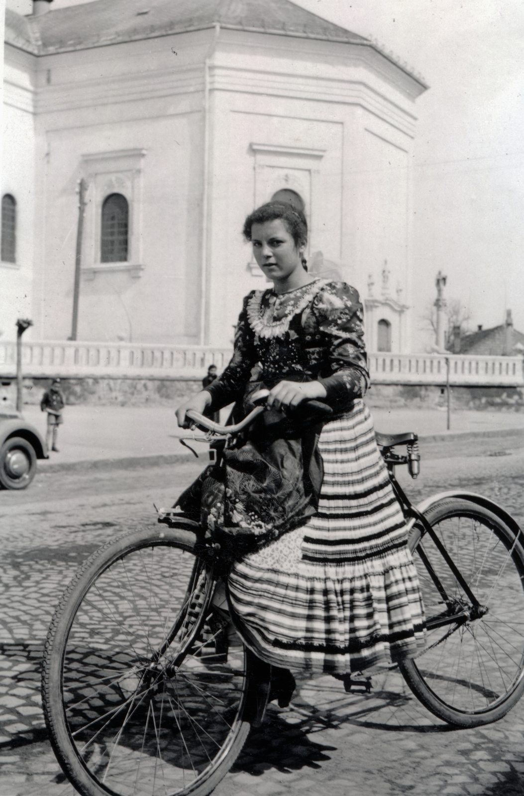 Hungary, Mezőkövesd, Mátyás király út, lány matyó népviseletben. Háttérben a Szent László-templom., 1939, Kőszegi Anna, bicycle, folk costume, skirt, Fortepan #151080