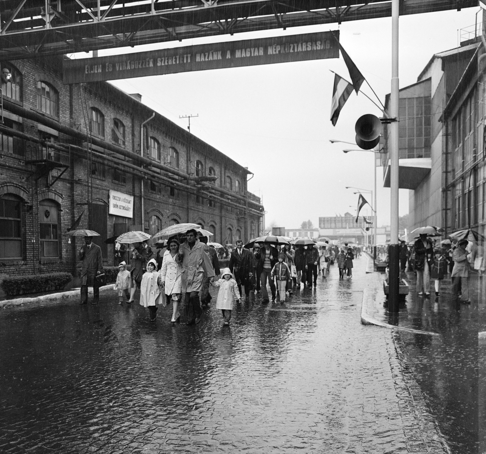 Magyarország, Budapest XXI., Csepel Vas- és Fémművek, Színesfém utca, szemben a főkapu a gyár felől nézve. MHSZ Gyermeknap a Nemzetközi Gyermekév tiszteletére., 1979, MHSZ, zászló, életkép, politikai dekoráció, esernyő, esőkabát, hangszóró, jelmondat, eső, Budapest, Fortepan #15818