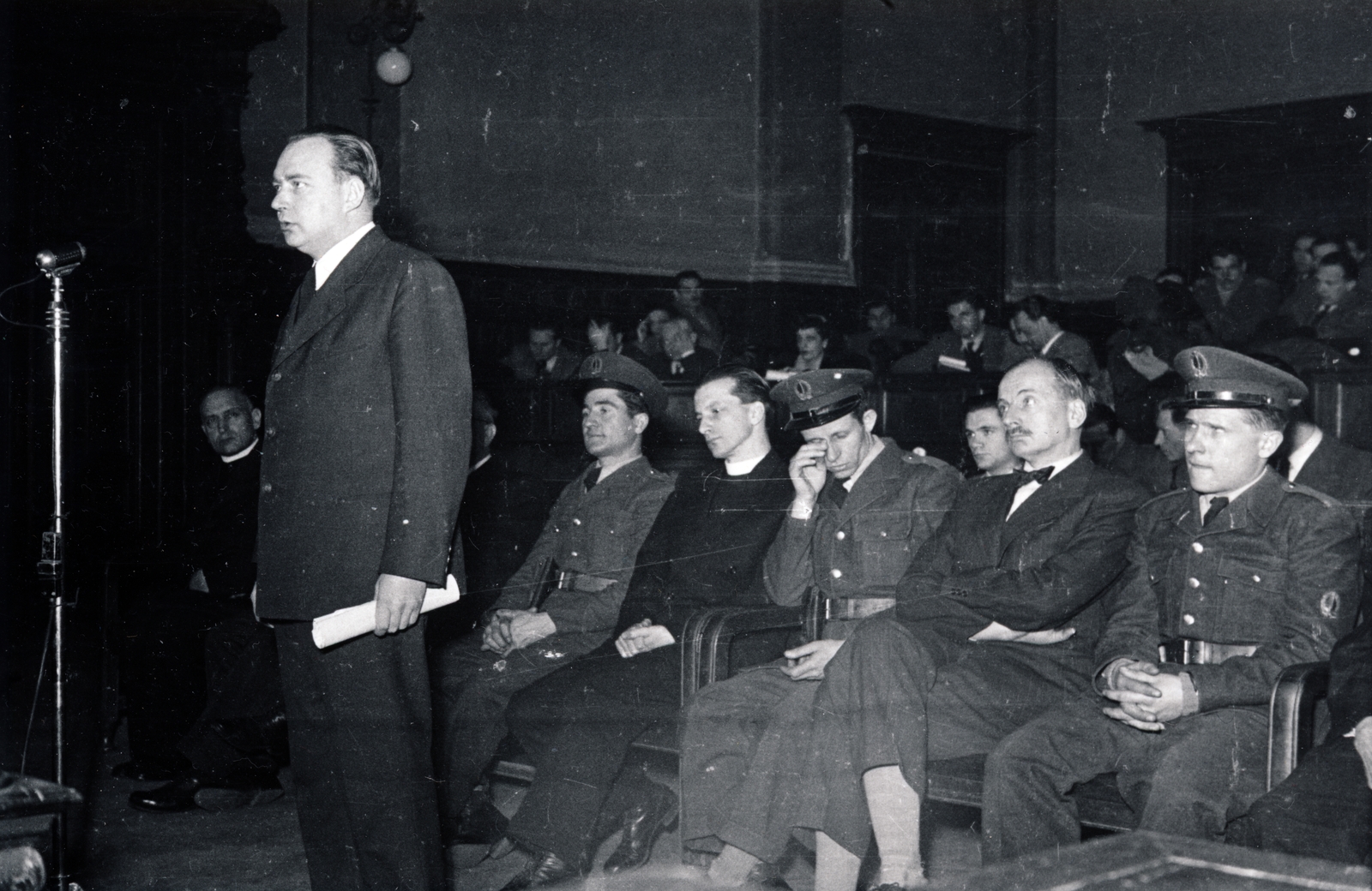 Magyarország, Budapest V., Markó utca 27., a Fővárosi Törvényszék épületének nagy esküdtszéki terme. A Budapesti Népbíróság különtanácsa által lefolytatott Mindszenty József bíboros, hercegprímás és vádlott társainak koncepciós pere 1949. február 3-5 között. A mikrofonnál dr. Ispánki Béla egyetemi lelkész, mögötte a vádlottak padján bal szélen Mindszenty József bíboros, hercegprímás, a fegyőrök között Zakar András érseki titkár és Esterházy Pál herceg., 1949, Fortepan/Album045, Bass Tibor, Budapest, bírósági tárgyalás, Fortepan #164866