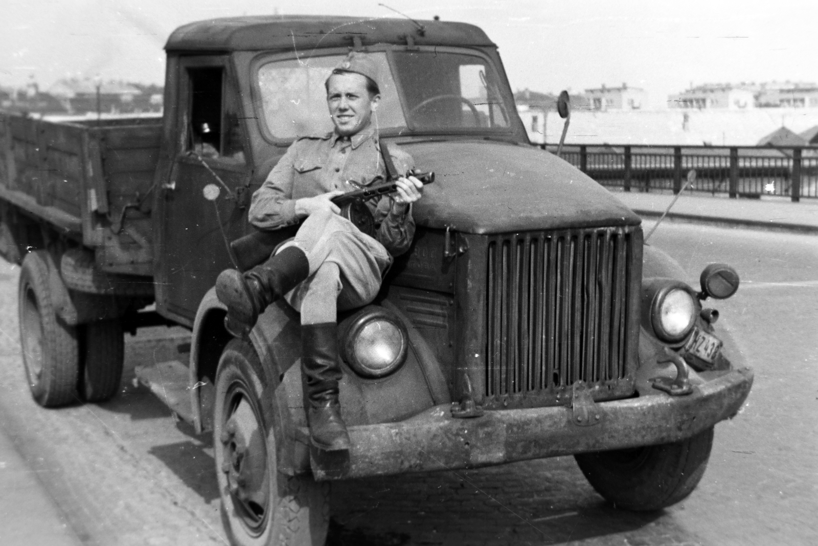 Hungary, Budapest XIII., Ferdinánd (Élmunkás) híd, jobbra a Nyugati pályaudvar területén lévő raktárak, a távolban a Lehel utcai "Élmunkás házak" látszanak., 1953, Tóth Tibor, Budapest, soldier, leg over leg, sitting on a car, commercial vehicle, weapon, PPS machine gun, GAZ-brand, Fortepan #185481