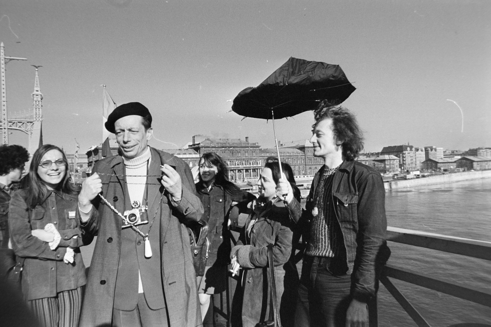 Hungary, Budapest IX., Szabadság híd, háttérben balra a Fővám (Dimitrov) tér, Marx Károly Közgazdaságtudományi Egyetem (később Corvinus Egyetem), jobbra a Közraktár utca. Harmath István fényképész mint a maga által, mindenek előtt magára vonatkoztatva alapított Világ Legnagyobb Marhája kitüntetés adományozója, a Bedobás kitalálója Dozvald János fotóművész, újságíró nyakába készül akasztani a kitüntetést jelképező WC-láncot. A felvétel az évente megtartott Bedobás / felesleges tárgyak Dunába dobása alkalmával készült., 1977, Gábor Viktor, Budapest, camera, umbrella, Fortepan #194207