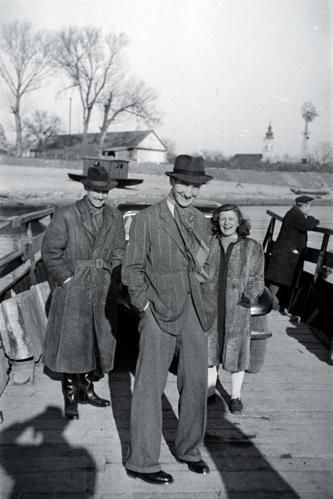 Hungary, Szob, a Szob-Pilismarót komp, háttérben a Luczenbacher hajóépítő műhely, a Szent László-templom, és egy szélkerék., 1941, Eisner Ferenc, ferry, hands in pockets, ship, three people, Fortepan #200303