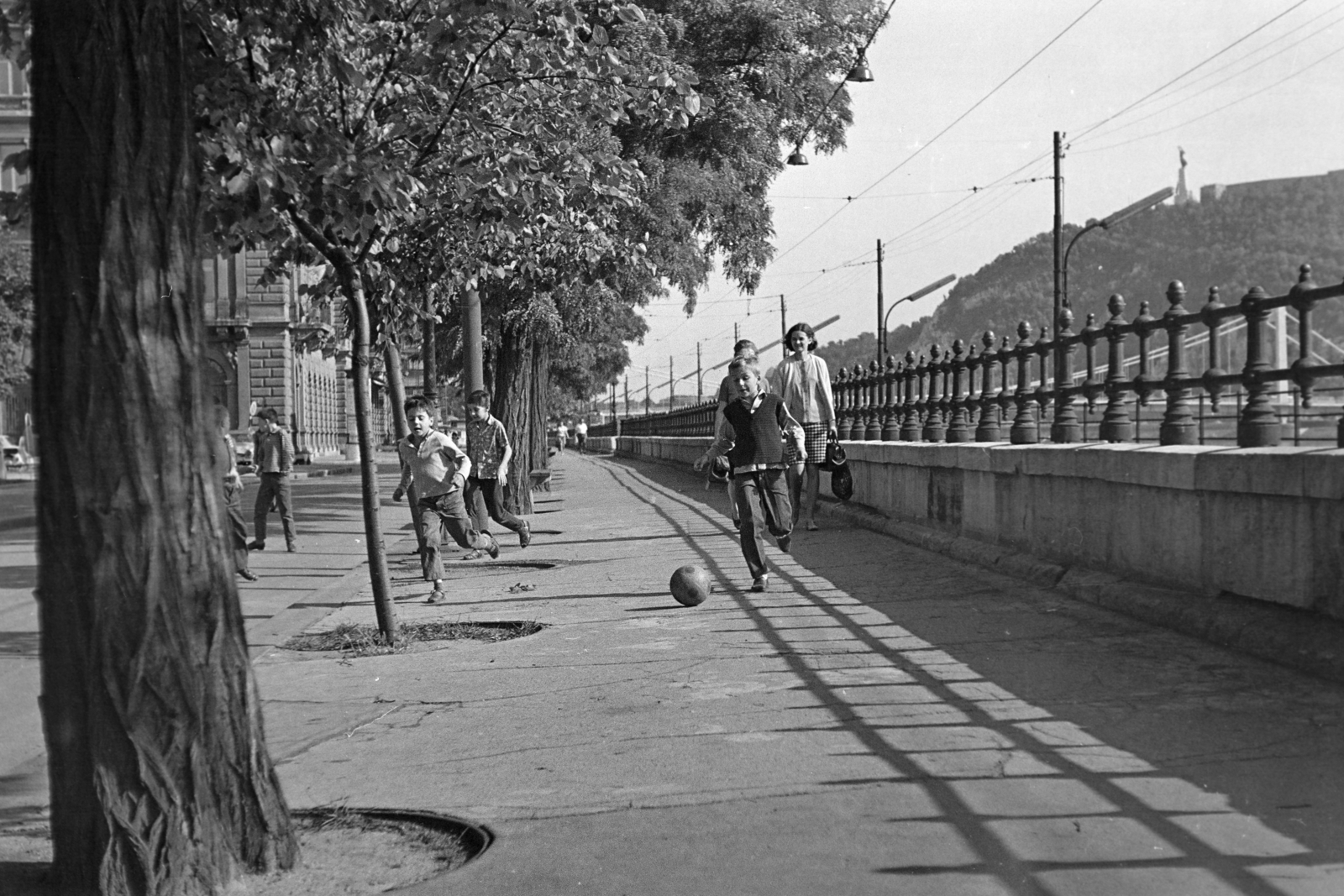 Magyarország, Budapest V., a Dunakorzó az Eötvös térnél, háttérben balra a Thonet-udvar a Wekerle Sándor (Szende Pál) utca sarkán, jobbra az Erzsébet híd és a Gellért-hegy., 1969, Kereki Sándor, Budapest, árnyék, fiúk, labda, Fortepan #204953