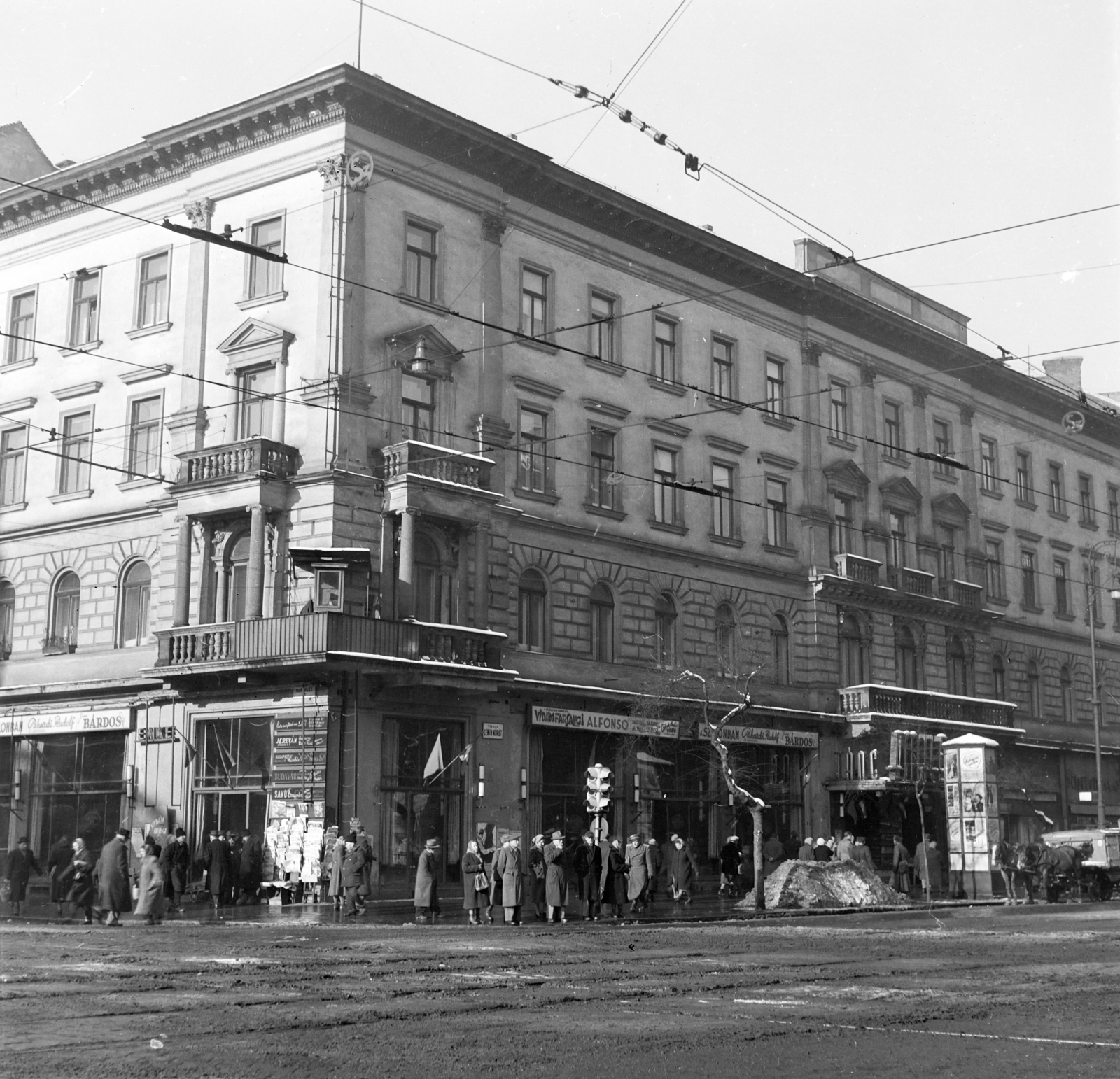 Hungary, Budapest VII., Rákóczi út - Erzsébet (Lenin) körút sarok. Érdekes a két I. emeleti erkélyt összekötő szerkezet, tetővel, a forgalomirányító rendőr számára., 1955, Kotnyek Antal, Budapest, photo aspect ratio: square, pedestrian, ashlar, balcony, ad pillar, newsstand, mud, catenary wire, street name sign, directing traffic, snow piles, intersection, Fortepan #210910