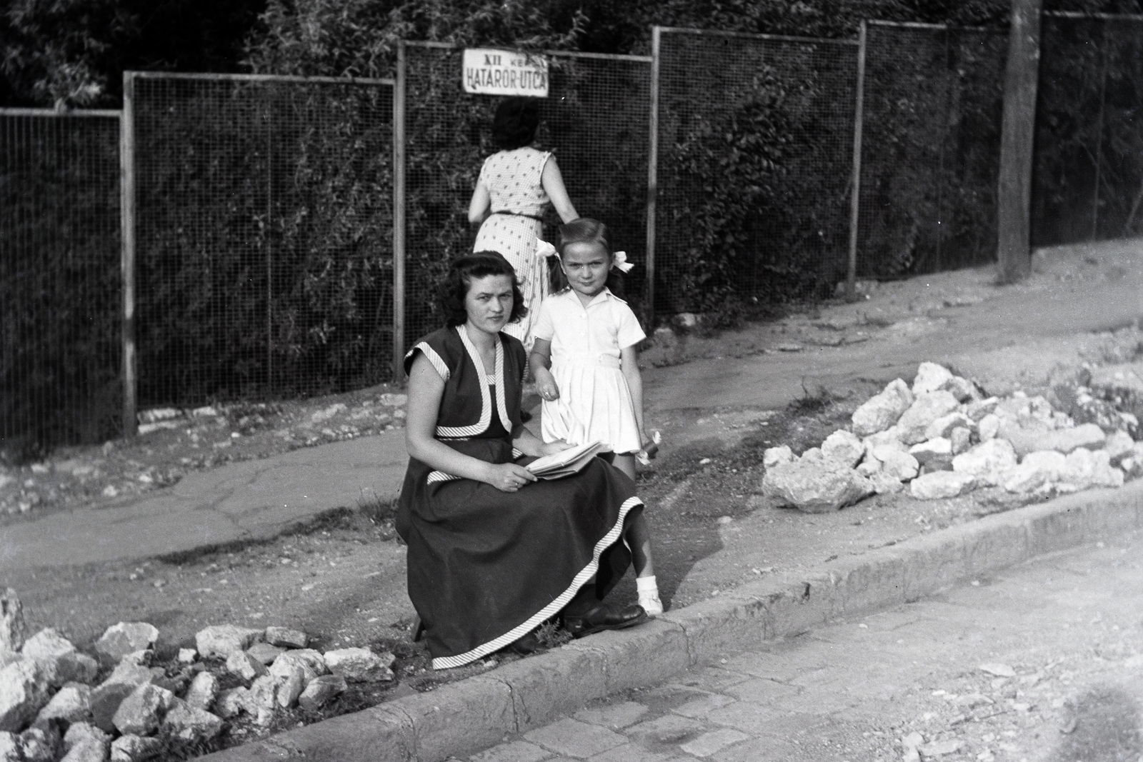 Hungary, Budapest XII., Határőr út (ekkor utca), a Gaál József utca 10. számú ház kerítése., 1951, Adelhardt Márta, Budapest, girl, ribbon, lady, street name sign, white dress, Fortepan #212393