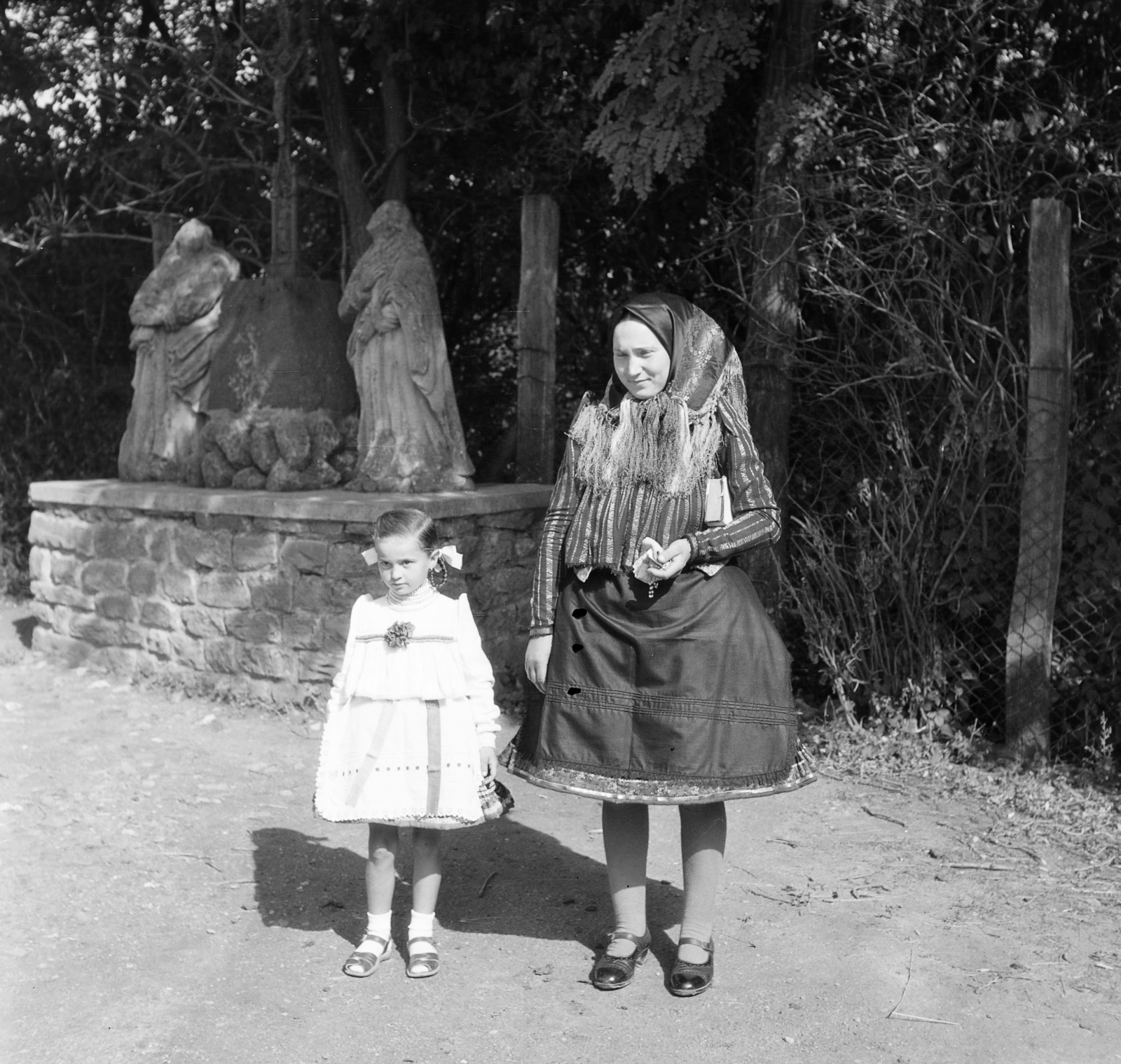 Hungary, Buják, a Tours-i Szent Márton-templom kertje. Szűz Mária és Szent János kőszobra között, a kálváriáról a XIX. század végén áthozott, 1859-ből származó vaskereszt., 1958, Adelhardt Márta, folk costume, Fortepan #212414