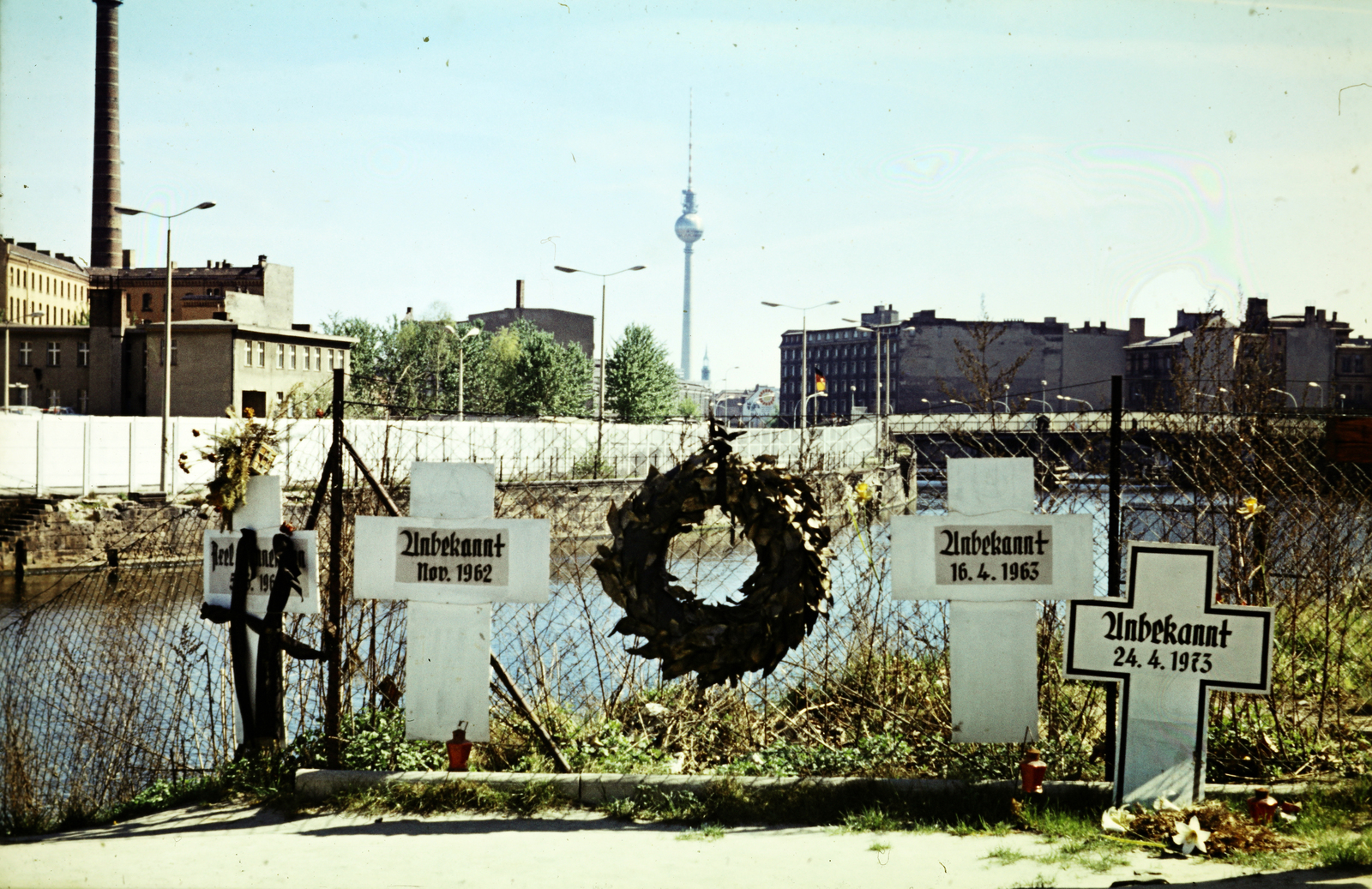 Németország, Berlin, Nyugat-Berlin, a Reichstagufer a Friedrich-Ebert-Platz (Ebertstrasse)-nál, Gedenkort "Weisse Kreuze" (Fehér Keresztek emlékhely). A Spree folyó túlsó partján a berlini fal, a háttérben a kép közepén a TV torony, alatta a St. Marienkirche tornya, a Spree folyó jobb partján félig takarva a fától (a TV torony aljánál) a Bhf Friedrichstrasse teteje, a híd a Marschallbrücke., 1973, Mezey András, színes, síremlék, Berlini fal, Nyugat-Berlin, Fortepan #219749