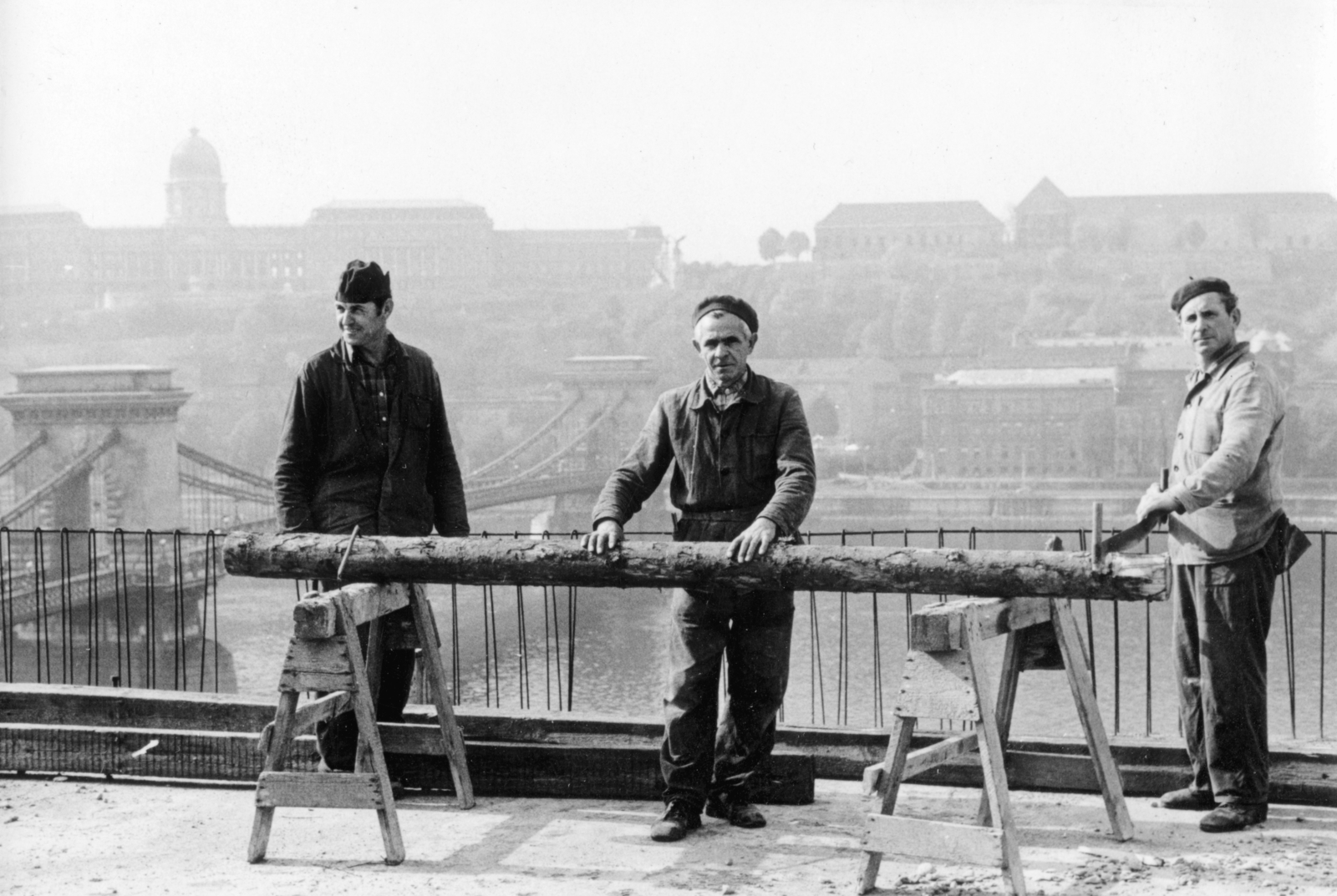 Hungary, Budapest V., Széchenyi István (Roosevelt) tér, a felvétel az állami külkereskedelmi vállalatok székházának tervezett irodaház (Spenótház) építkezésén készült. Háttérben a Széchenyi Lánchíd és a budai Vár., 1974, Középületépítő Vállalat - Kreszán Albert - Koczka András - Kemecsei József, Budapest, worker, saw-buck, saw, trunk, Best of, Fortepan #227728