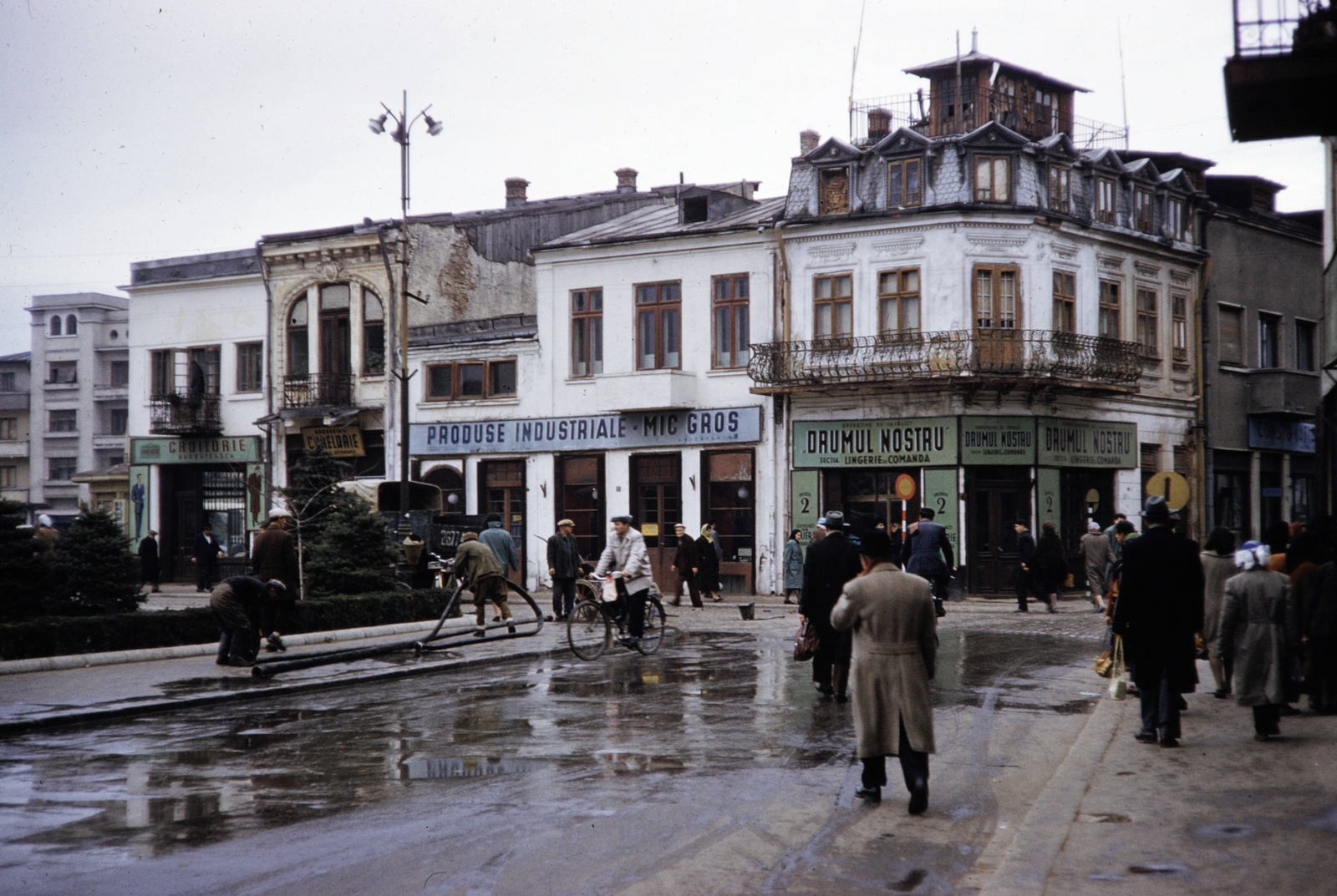 Románia, Ploiesti, Strada Romană, keresztbe a Strada Lipscani. A szemben lévő épületeket az 1977-es földrengés után lebontották, ma e helyen a Piața Ion Luca Caragiale található. Bal szélen a háttérben a mai is álló Strada Emile Zola 2., 1960, UWM Libraries, Harrison Forman, színes, üzletportál, utcakép, kerékpár, eső, román felirat, Fortepan #260492
