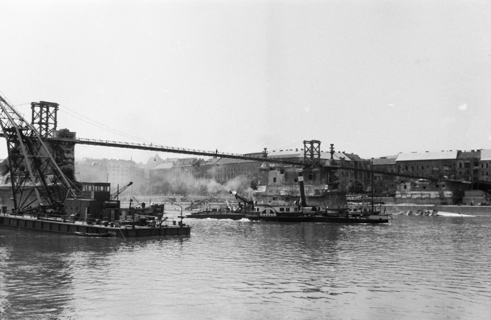 Magyarország, Margitsziget, Budapest II., a lerombolt Margit híd újjáépítésén dolgozó József Attila úszódaru, a felvétel a roncsok kiemelésekor készült. Fent a híd pillérein épült, fajármokra szerelt, közművezetékeket is hordozó kábelhíd látható. A Dunán, a megtisztított mederszakaszon, a Kazán oldalkerekes vontató gőzhajó halad., 1947, Vízkelety László, Budapest, hídroncs, hajódaru, gőzhajó, Fortepan #266258