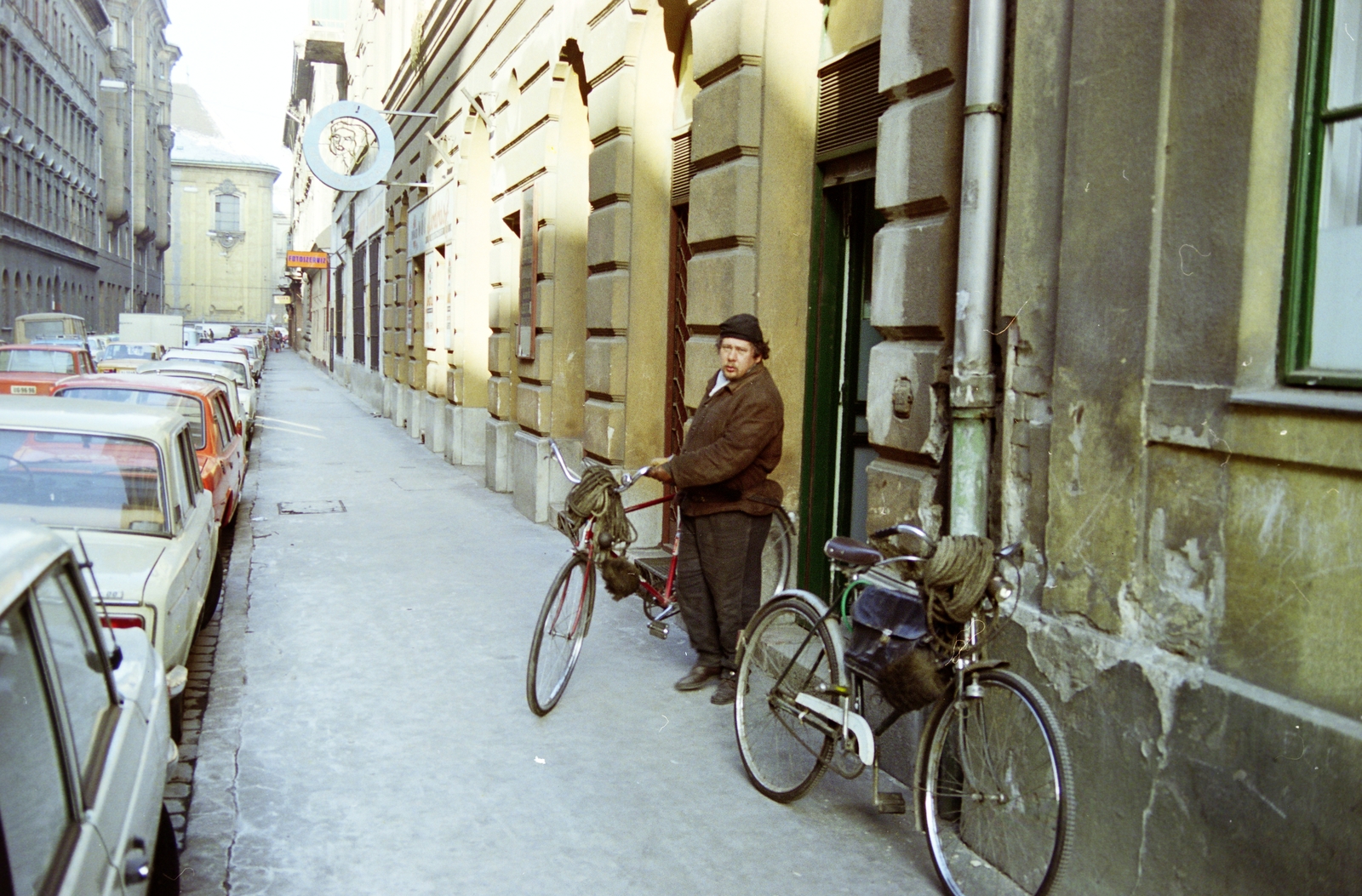 Magyarország, Budapest VII., Akácfa utca, munkába induló kéményseprő, az 57. számú házban a Fővárosi Kéményseprő és Tüzeléstechnikai Szolgáltató Vállalat irodája és öltözője előtt. Távolban a Király (Majakovszkij) utcánál az Avilai nagy Szent Teréz-templom (Terézvárosi templom) látható., 1983, Kanyó Béla, Budapest, kéményseprő, kerékpár, színes, Fortepan #269134