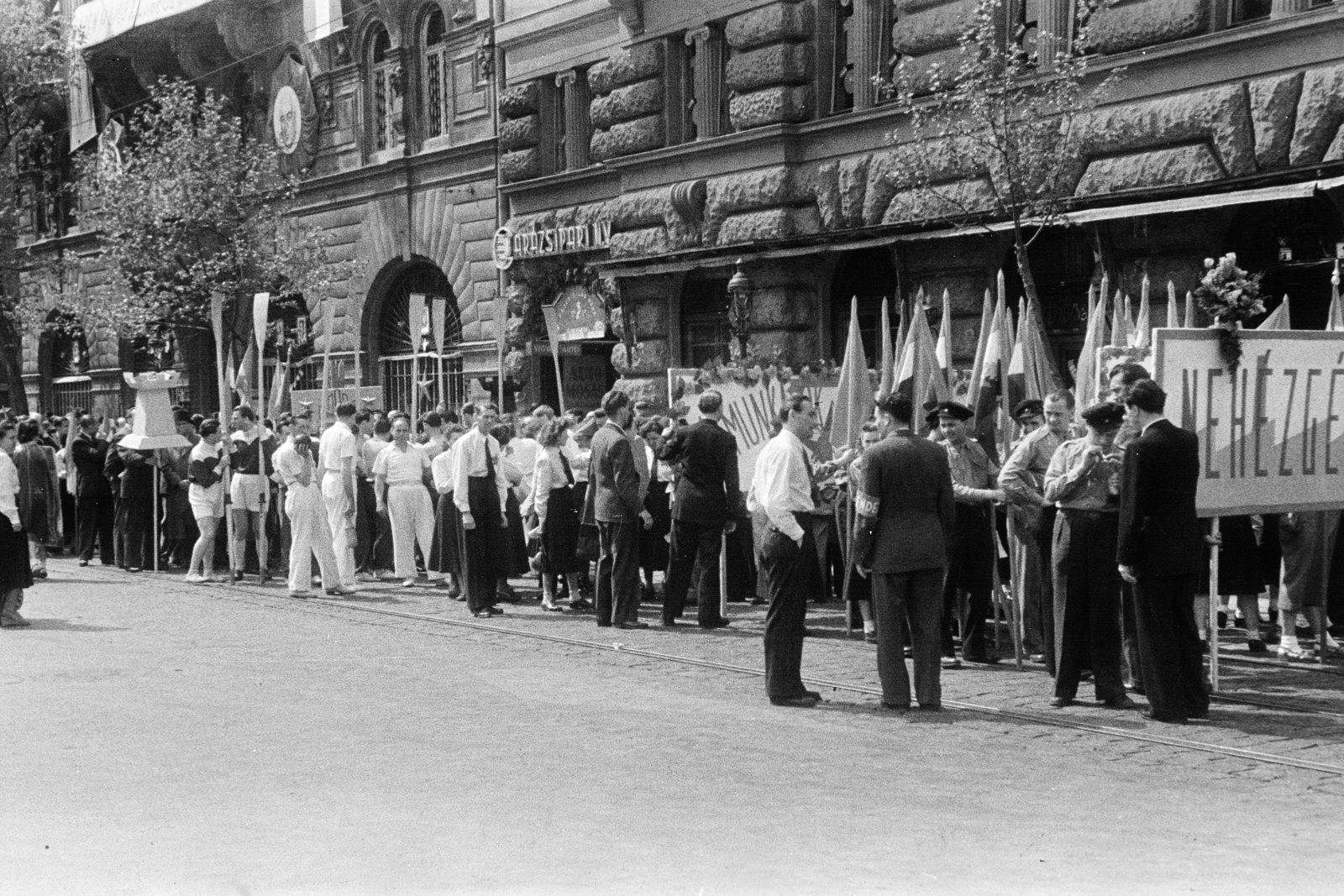 Magyarország, Budapest V., Vértanúk tere (Ságvári tér), felvonulás résztvevői. A felvétel a Kossuth Lajos tér sarkán álló ház előtt készült, balra a Nádor utca 36-os számú épület., 1950, Kern család, Budapest, várakozás, felvonulás, transzparens, zászló, Fortepan #273328