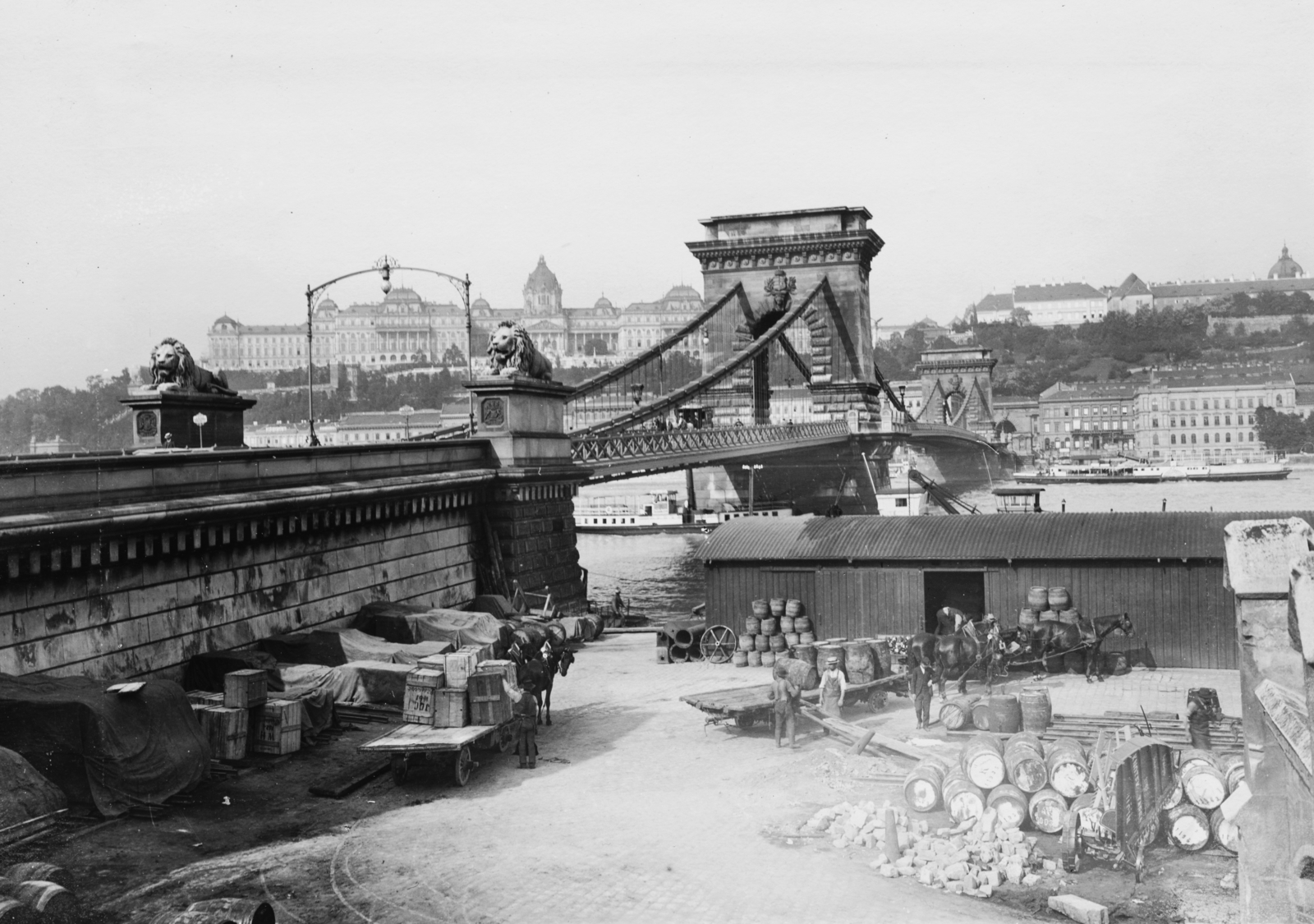 Hungary, Budapest V., pesti alsó (Rudolf) rakpart a Széchenyi Lánchíd hídfőjénél, a teherhajó kikötő egyik raktára. Balra a háttérben a Királyi Palota (később Budavári Palota) látható., 1907, Deutsche Fotothek / Brück und Sohn, Best of, barrel, chest, coach, stone lion, William Tierney Clark-design, suspension bridge, Budapest, Fortepan #277807