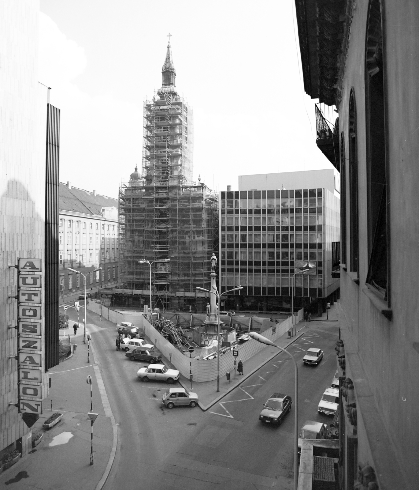 Magyarország, Budapest V., Szervita (Martinelli) tér a Kristóf tér irányából a Városház utca felé nézve, mellette a felállványozott Belvárosi Szent Anna-templom (szervita templom). Jobbra a Belvárosi Távbeszélő Központ a Petőfi Sándor utcánál. Középen a Mária oszlop. Bal szélen az Országos Műszaki Fejlesztési Bizottság (OMFB) székháza földszintjén, a Merkur Személygépkocsi Értékesítő Vállalat szalonja., 1981, Artfókusz, Fábián József, Budapest, Fortepan #280208