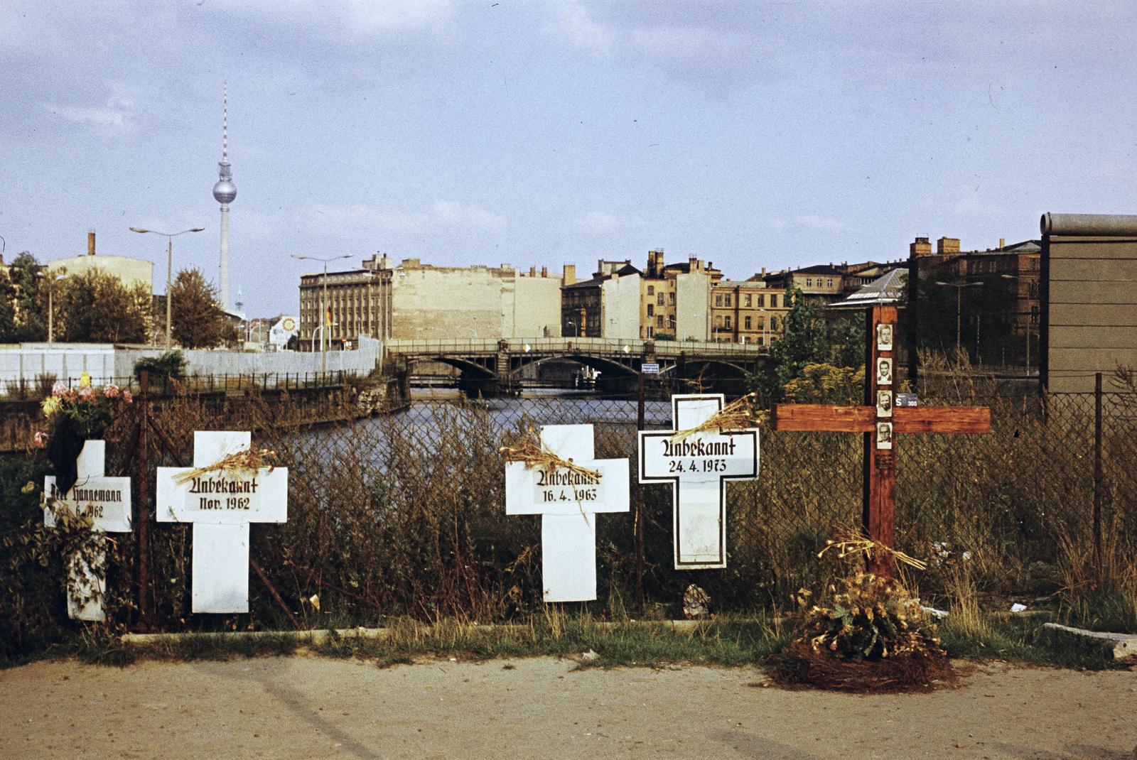Germany, Berlin, Nyugat-Berlin, a Reichstagufer a Friedrich-Ebert-Platz (Ebertstrasse)-nál, Gedenkort "Weisse Kreuze" (Fehér Keresztek emlékhely). A Spree folyó túlsó partján a berlini fal, a háttérben a kép közepén a TV torony, alatta a St. Marienkirche tornya, a Spree folyó jobb partján félig takarva a fától (a TV torony aljánál) a Bhf Friedrichstrasse teteje, a híd a Marschallbrücke., 1975, Szomolányi József, Best of, colorful, cross, headstone, chain-link fence, bridge, Fortepan #302818