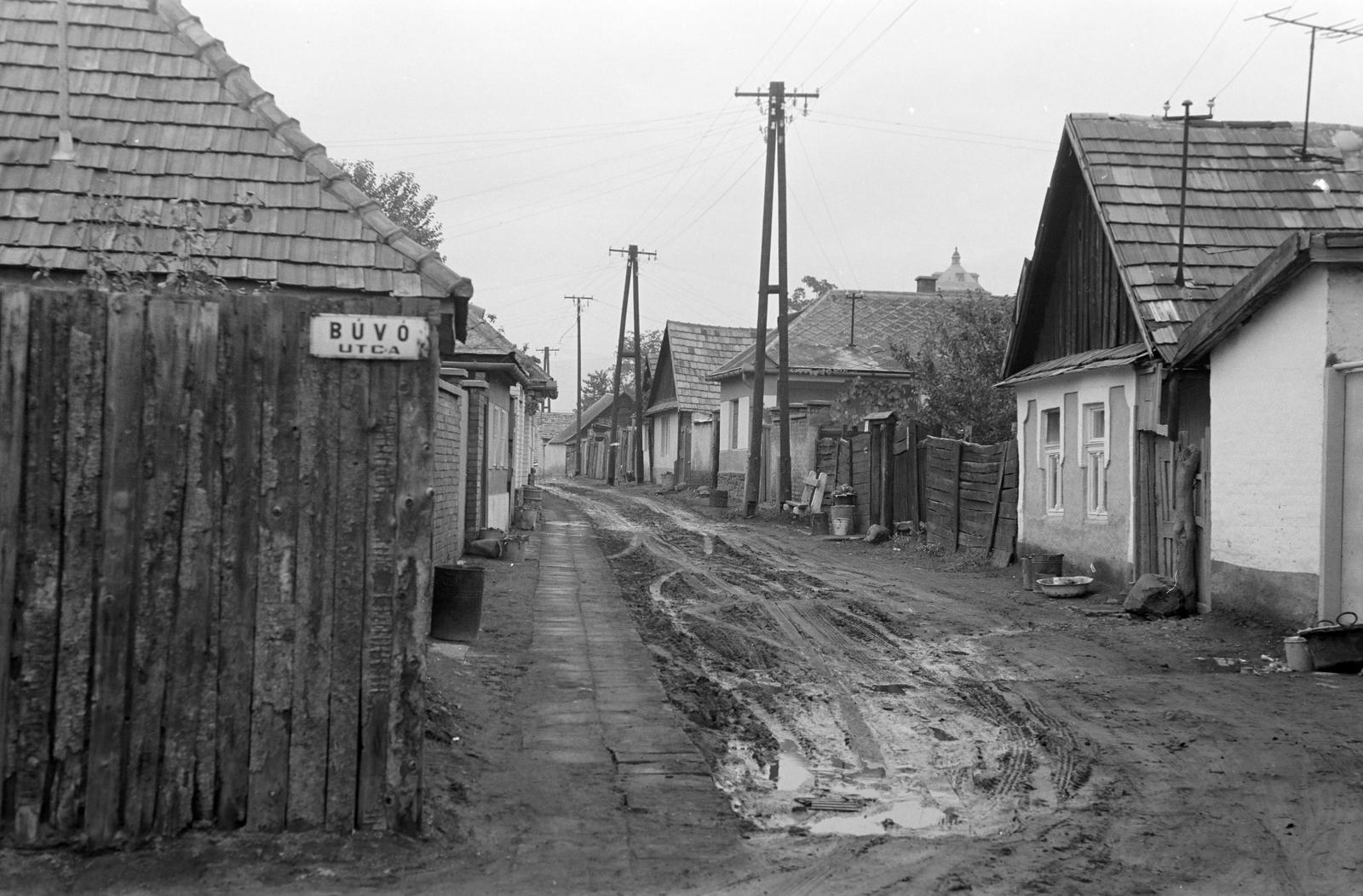 Hungary, Gyöngyös, Búvó utca - Verő utca sarok. Jobbra a háztetők felett a Petőfi Sándor utcai Víztorony., 1966, Gazda Anikó, street view, pylon, dirt road, house, street name sign, trash can, bench, mud, Fortepan #307945