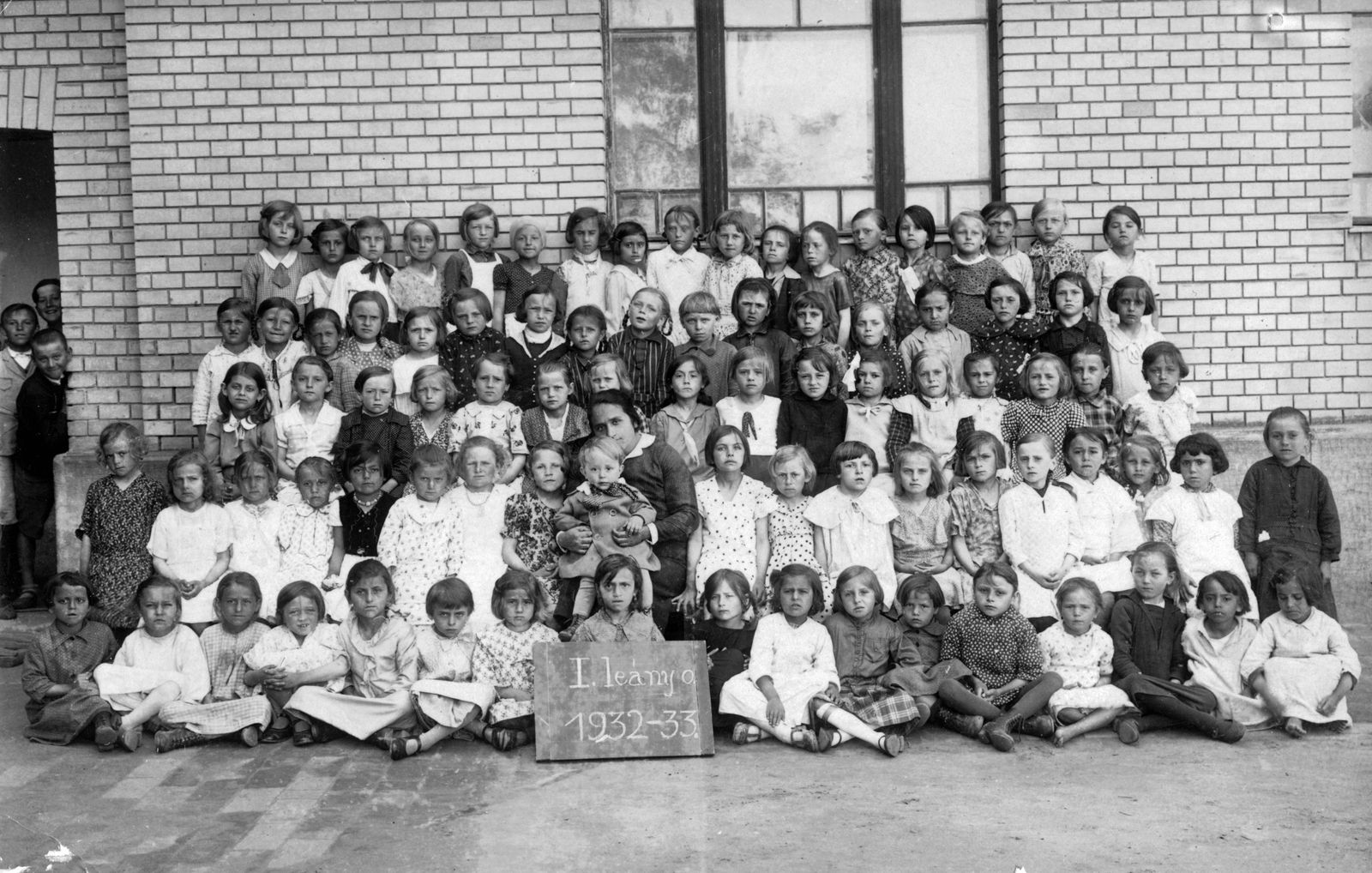 Hungary, Kunmadaras, Református Elemi Népiskola. 1932-33-as tanév, csoportkép az I/a osztályról., 1933, Anderle Lajosné, label, class photo, girl, girls' class, kids, teacher, board, peeking, cross-legged sitting, Fortepan #31415