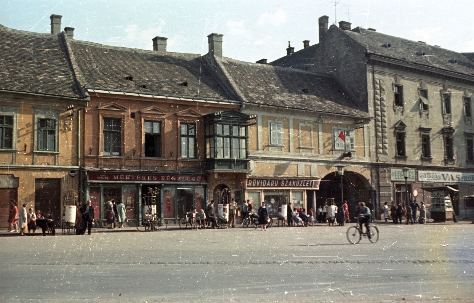 Hungary, Pápa, Fő tér, jobbra az átjáró a Kossuth utcára., 1961, Nagy Gyula, bicycle, street furniture, colorful, street view, genre painting, ad pillar, lamp post, store display, bench, newsstand, shoemaker, haberdashery, candy store, hardware store, Fortepan #51005