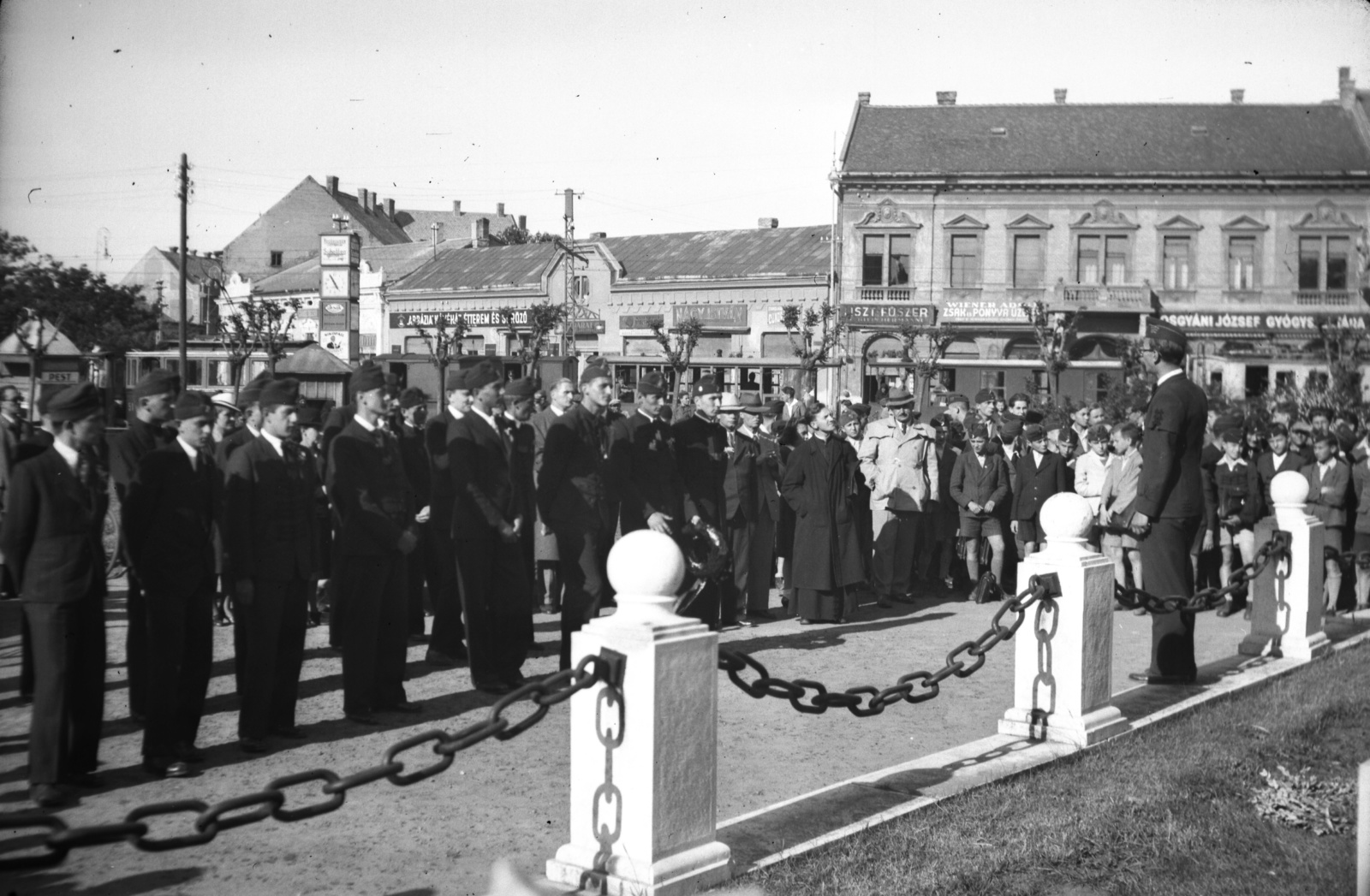 Hungary, Nyíregyháza, Hősök tere (Bessenyei tér), ünnepség a Hősök szobra előtt. Háttérben az NyVKV (Nyíregyházavidéki Kisvasutak) szerelvénye és Osgyáni József gyógyszertára (korábban: Korányi-patika)., 1938, Zsanda Zsolt, sign-board, watch, street view, festive, narrow-gauge railway, Fortepan #58733