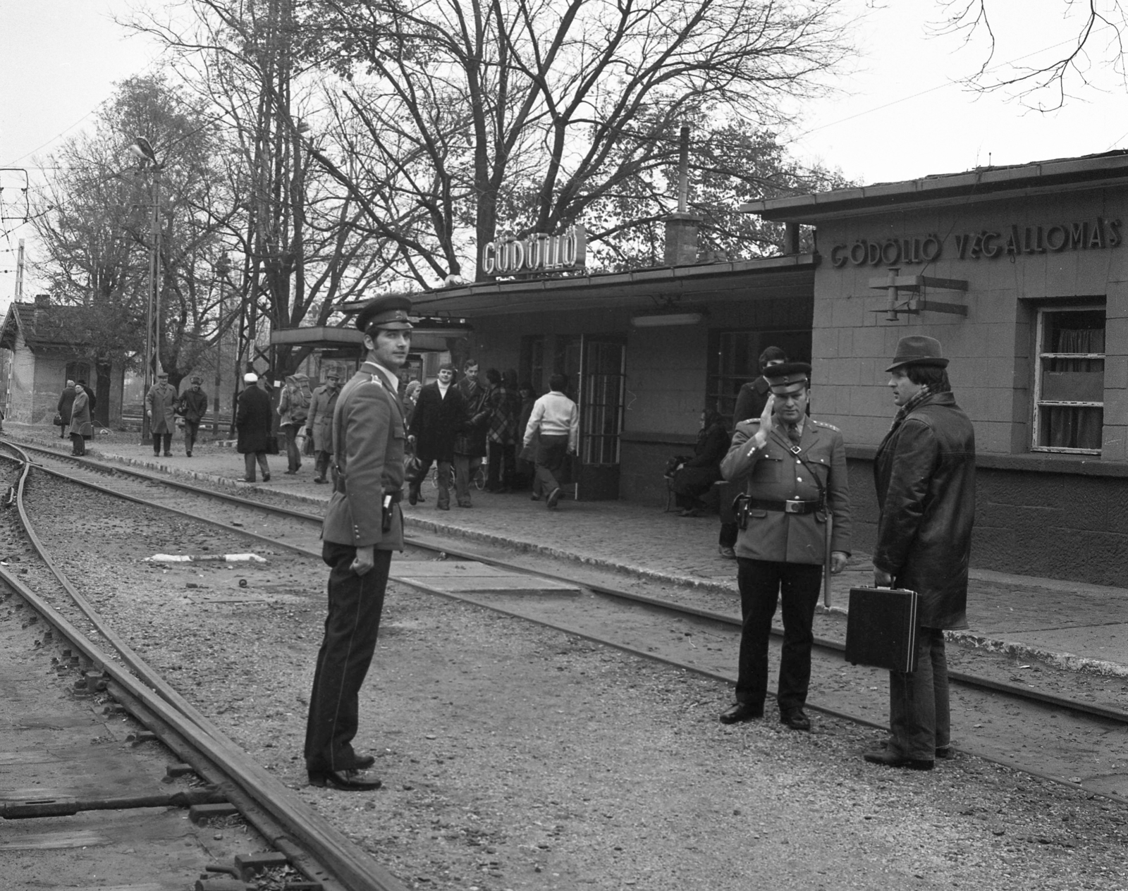 Hungary, Gödöllő, HÉV végállomás., 1981, Magyar Rendőr, hat, uniform, cop, man, salute, train station, commuter train, peaked cap, passenger, rails, neon sign, place-name signs, attaché case, Fortepan #66692