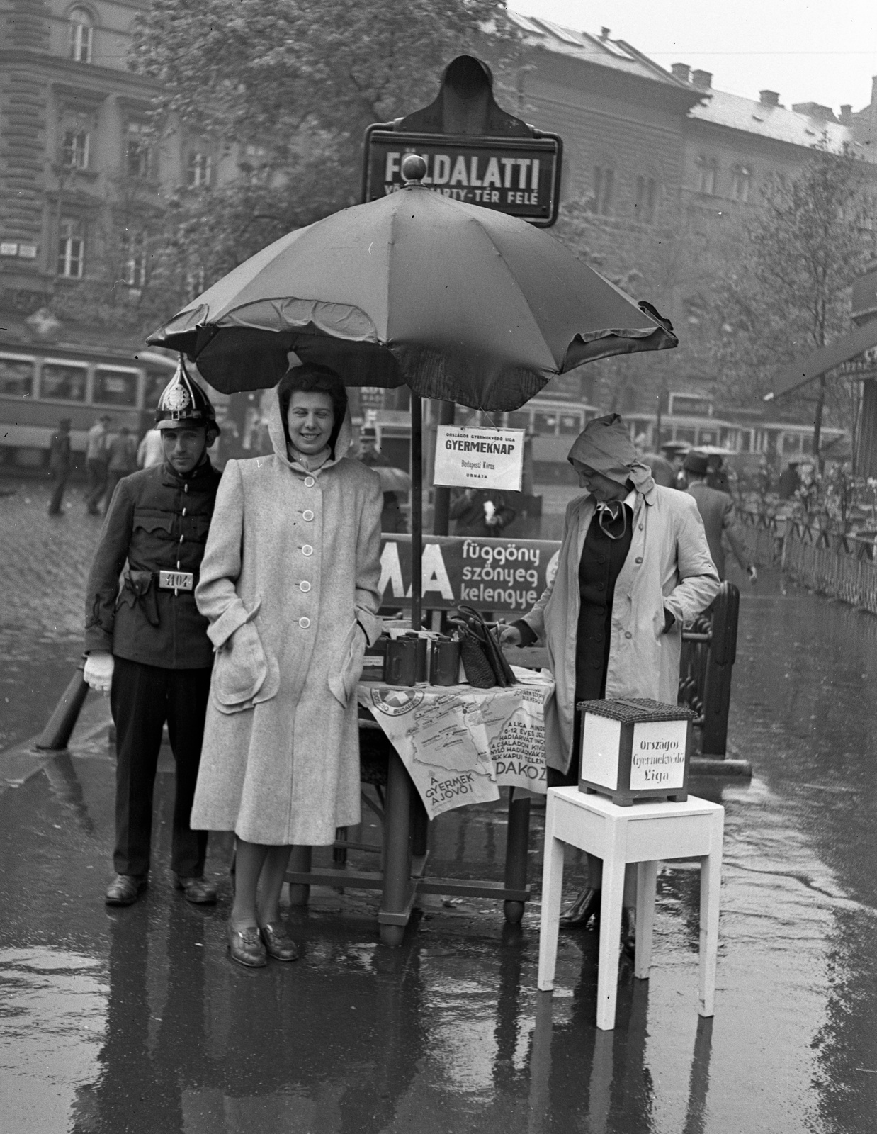 Hungary, Budapest VI., Oktogon, a Budapesti Kórus tagjai adományokat gyűjtenek az Országos Gyermekvédő Liga számára az Abbázia Kávéház előtt., 1943, Lissák Tivadar, cop, charity, underground railway, money-box, Budapest, kitchen stool, rain, raincoat, Fortepan #72078