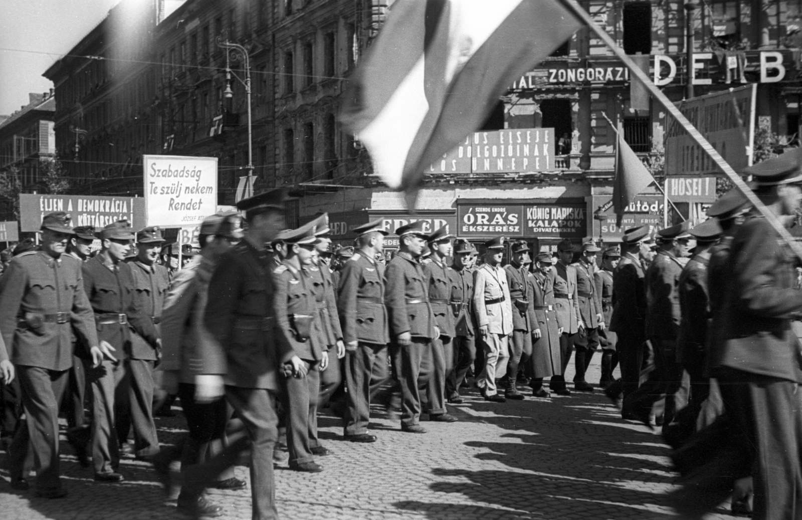 Magyarország, Budapest VI., Oktogon, a május 1-i ünnepség felvonulói a Teréz körútról kanyarodnak az Andrássy út felé., 1946, Berkó Pál, egyenruha, zászló, cégtábla, utcakép, életkép, rendőr, politikai dekoráció, felvonulás, lámpaoszlop, kockakő, órás, pékség, rendőrség, kalapos, Budapest, Fortepan #78621