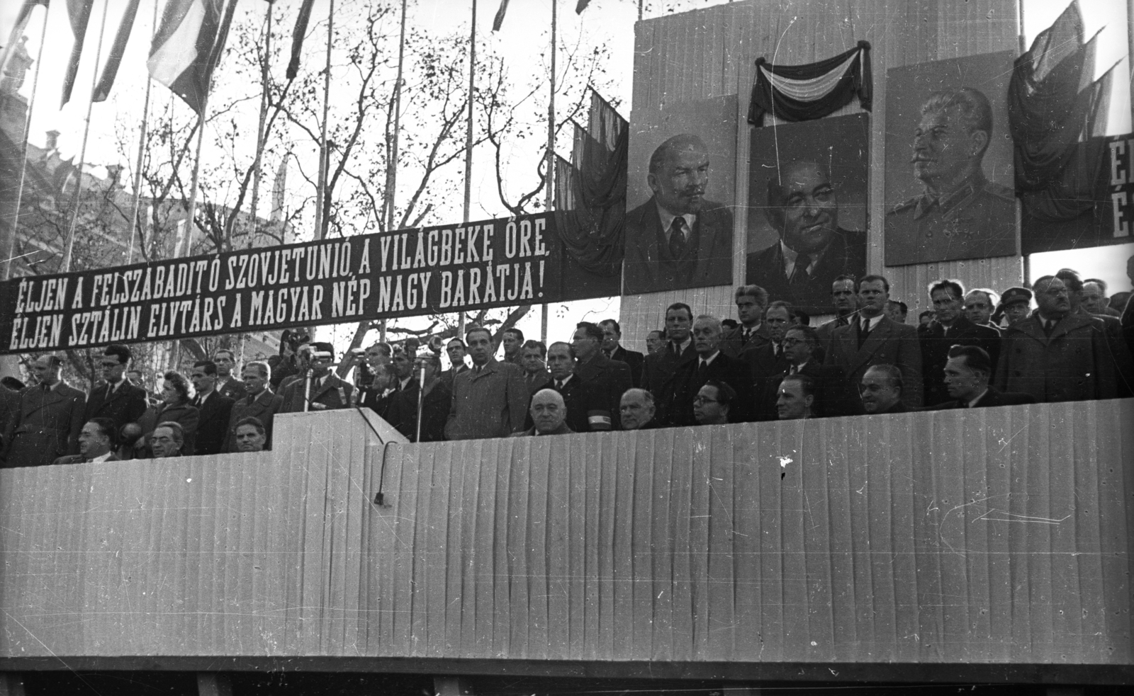 Hungary, Budapest V., Széchenyi István (Roosevelt) tér, balra a mai Belügyminisztérium épülete látszik. Az újjáépített Széchenyi Lánchíd ünnepségének tribünjén az első sorában ülnek, balról: Vas Zoltán, Gerő Ernő, Dobi István, Rákosi Mátyás, Bebrits Lajos, Kállai Gyula, Kádár János, Ries István, Zsofinyecz Mihály., 1949, UVATERV, celebrity, personality cult, Lenin-portrayal, Mátyás Rákosi-portrayal, Joseph Stalin portrayal, Budapest, politician, Fortepan #80344