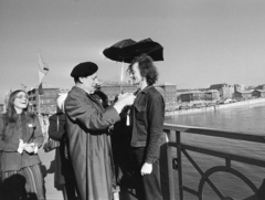 Magyarország, Budapest IX., Szabadság híd, háttérben balra a Fővám (Dimitrov) tér, Marx Károly Közgazdaságtudományi Egyetem (később Corvinus Egyetem), jobbra a Közraktár utca. Harmath István fényképész mint a maga által, mindenek előtt magára vonatkoztatva alapított Világ Legnagyobb Marhája kitüntetés adományozója, a Bedobás kitalálója Dozvald János fotóművész, újságíró nyakába akasztja a kitüntetést jelképező WC-láncot. A felvétel az évente megtartott Bedobás / felesleges tárgyak Dunába dobása alkalmával készült., 1977, Gábor Viktor, Budapest, Fortepan #194206