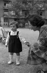 Magyarország, Budapest XI., Kosztolányi Dezső téri park, háttérben a 9. számú ház., 1954, Martin Kornél, Budapest, babakocsi, anya, kézenfogva, kislány, hajviselet, kötényruha, kockás ruha, Fortepan #196728