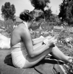 Hungary, Balatonfűzfő, a Petrik Lajos Vegyipari Technikum egyik diáklánya nyári táborban. Háttérben balra a vitorlás klub épülete látszik., 1958, Ungváry Rudolf, bathing suit, girl, hairdo, newspaper, reading, Fortepan #254932