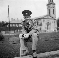 Hungary, Budapest XIII., a Kassák Lajos utca (Fóti út) - Taksony utca - Tüzér utca - Lőportár utca határolta park, háttérben a Kassák Lajos utca (Fóti út) és az angyalföldi evangélikus templom., 1951, Ungváry Rudolf, Budapest, peaked cap, children's railway personnel, teenager, boy, sitting on backrest of a bench, Fortepan #255040