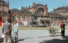 Németország, Schwerin, főpályaudvar, előtérben a Megmenekült hajótörött (Rettung aus Seenot, Hugo Berwald, 1911) szőkőkút., 1962, Fortepan, utcabútor, színes, zászló, utcakép, életkép, politikai dekoráció, pihenés, NDK, lámpaoszlop, pad, pályaudvar, turistabot, jelmondat, kézitáska, Ernst Moeller-terv, Fortepan #44289