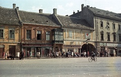 Magyarország, Pápa, Fő tér, jobbra az átjáró a Kossuth utcára., 1961, Nagy Gyula, kerékpár, utcabútor, színes, utcakép, életkép, hirdetőoszlop, lámpaoszlop, üzletportál, pad, újságárus, cipész, rövidáru, Édességbolt, háztartási bolt, Fortepan #51005